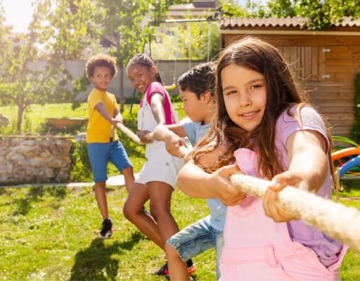 Tauziehen im Garten Ein sonniges Outdoor-Foto zeigt vier Kinder, die im grünen Garten Tauziehen spielen. Im Vordergrund zieht ein Mädchen in rosa Shirt kräftig am Seil, während drei weitere Kinder im Hintergrund mitlachen und dagegenhalten – fröhliche Bewegung und Teamgeist.